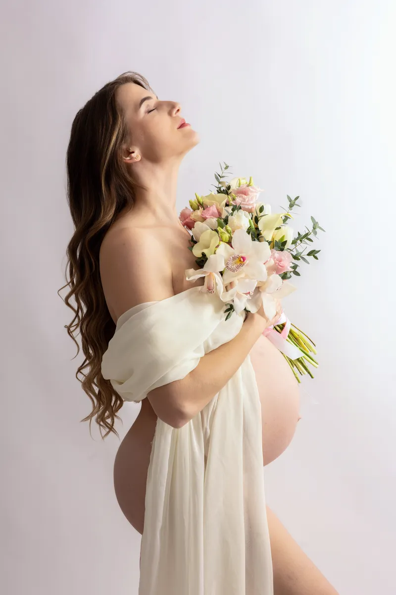 Eine schwangere Frau im Profil hält einen Blumenstrauß in einem Studio.